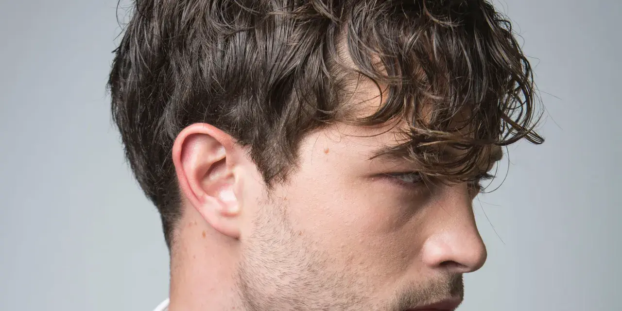 Perfil de hombre con cabello castaño ondulado y barba incipiente. Su corte de pelo es un rapado al 3 en los laterales.